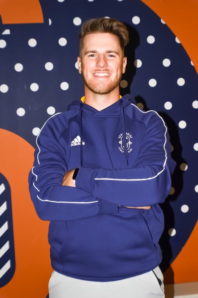 Smiling person in blue hoodie with crossed arms standing in front of a polka-dot wall.