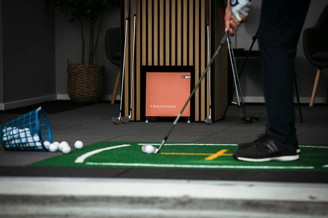 Indoor golf setup with Trackman device and golfer preparing to putt on green mat.