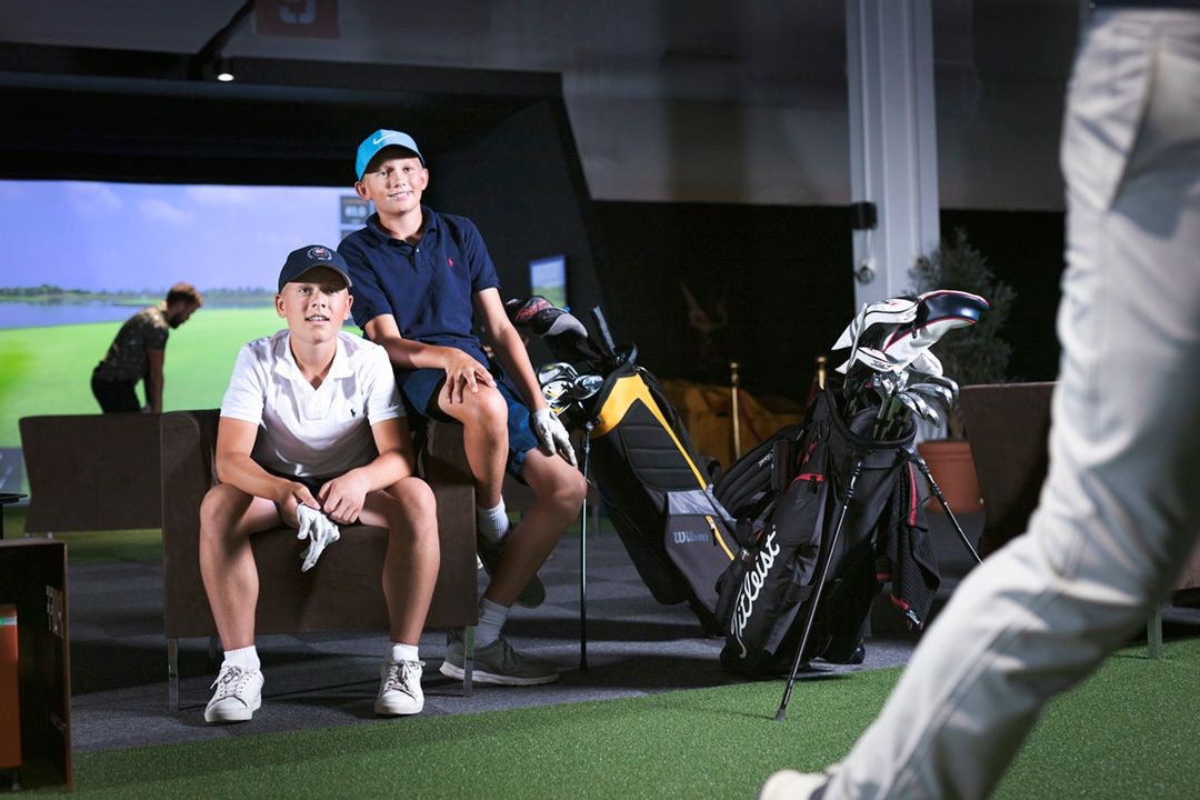 Two young boys in golf attire sitting indoors with golf bags and equipment nearby.
